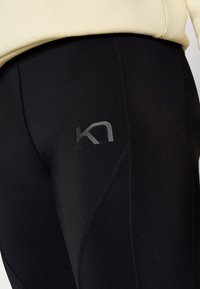 Primo piano di leggings sportivi neri con un piccolo logo grigio sulla coscia, indossati con una maglietta beige, mettendo in evidenza la texture del tessuto e i dettagli delle cuciture.