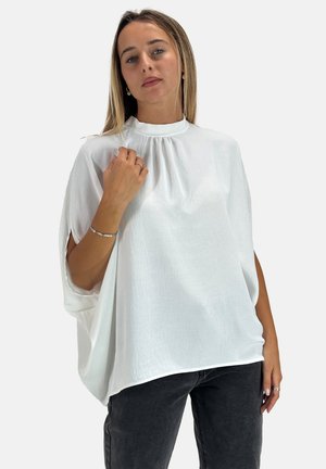 Hvid, løstsiddende bluse med høj hals og subtil tekstur, med åbne sider og en buet kant. Kombineret med mørkegrå bukser.