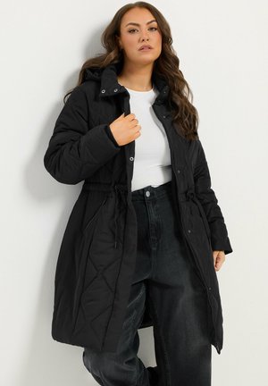Femme portant une longue veste noire matelassée avec capuche, chemise blanche et jean noir taille haute, debout contre un mur blanc.
