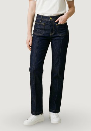 Persona che indossa jeans blu scuro a gamba dritta con tasche applicate frontali, sneakers bianche e una maglietta bianca a maniche corte, mano in tasca.