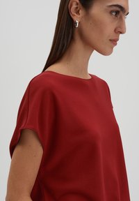 Rotes, kurzärmeliges Oberteil mit glatter Textur und rundem Halsausschnitt. Verfügt über dezente Schulterdetails und besteht aus einem leichten Stoff.