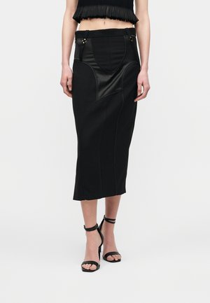 Kvinde iført en sort midi pencilnederdel med skinnende stofpaneler, kombineret med sorte ankelrem høje hæle sandaler og en cropped top.