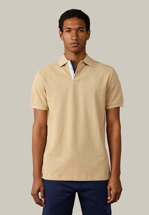 Junger Mann, der nach vorne schaut, trägt ein beige-farbenes kurzärmeliges Polohemd mit Kontrastbesatz und dunkelblaue Hose.