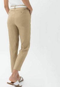 Pantaloni beige, su misura, con taglio dritto, dotati di tasche posteriori e una texture liscia. Abbinati a sandali bianchi.