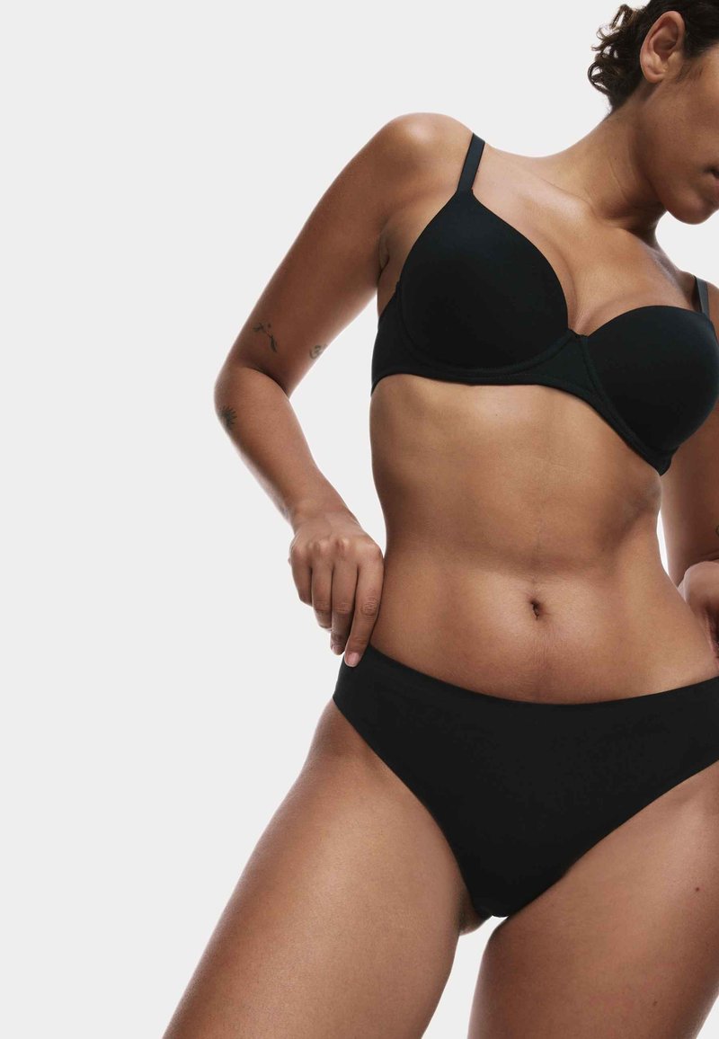 Donna che sistema un set di reggiseno e mutande nere, mostrando il torso superiore e i fianchi su uno sfondo neutro.