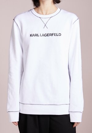 Witte lange mouwen trui met zwarte tekst "KARL LAGERFELD" op de borst en contrasterende stiksels, gedragen met zwarte broek.