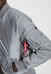 Gray bomber jacket with ribbed cuffs, featuring a zip pocket on the sleeve, a keychain loop, and smooth, shiny fabric texture.