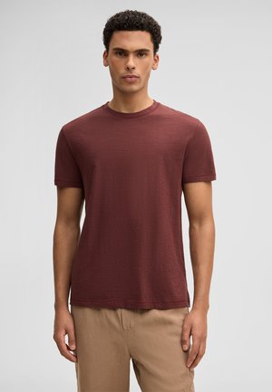 Man met kort krullend haar, gekleed in een effen bordeauxrood T-shirt met korte mouwen en beige broek, tegen een lichtgrijze achtergrond.