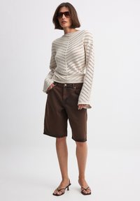 Suéter de punto a rayas en color beige claro, con cuello barco y mangas largas acampanadas; pantalones cortos de mezclilla marrones, hasta la rodilla, con dobladillos deshilachados. Sandalias negras.