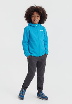 Παιδί με σγουρά μαλλιά που χαμογελά, φοράει μπλε ζακέτα με φερμουάρ, σκούρο γκρι παντελόνι και μπλε-γκρι αθλητικά παπούτσια, στέκεται με τα χέρια στις τσέπες.