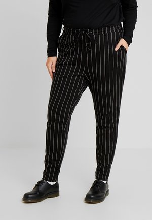 Person trägt schwarze Nadelstreifenhose und schwarze Schuhe, mit einer Hand in der Tasche und einem schwarzen langärmeligen Oberteil.