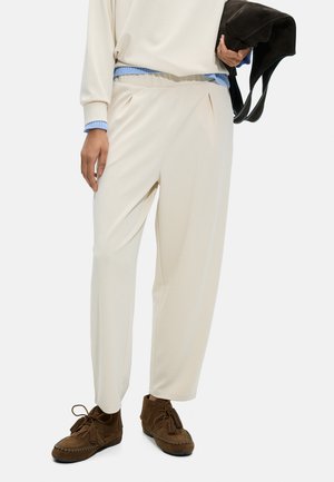 Osoba nosząca kremowe, luźne spodnie, jasnoniebieski sweter pod białym topem, trzymająca czarną torebkę i brązowe mokasyny.