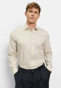 Beiges Langarmhemd mit subtiler Struktur, Knopfleiste vorne und spitzem Kragen, kombiniert mit dunkler Hose, vor weißem Hintergrund.