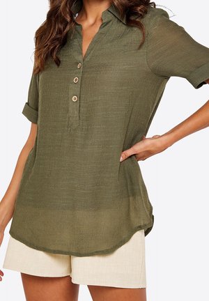 Femme portant un chemisier boutonné à manches courtes vert olive avec les poignets retroussés et un short beige, debout, une main sur la hanche.