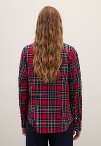 Red plaid shirt with green and yellow accents, long sleeves, and button cuffs. Features a slightly curved hem and soft, textured fabric.