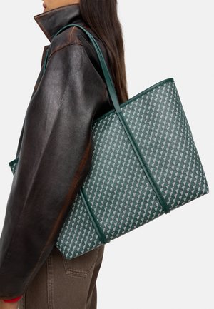 Zielona torba na ramię w wzory z teksturowanym wykończeniem, uzupełniona jednolitymi, zielonymi ramiączkami. Nosa na tle skórzanej kurtki z podniesionym kołnierzem.