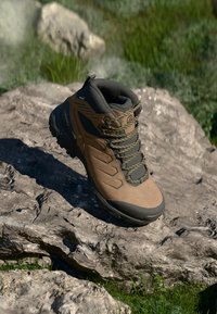 Bota de senderismo marrón con parte superior sintética, suela de goma, cuello acolchado y acentos en negro. Cuenta con puntera reforzada y superficie texturizada.