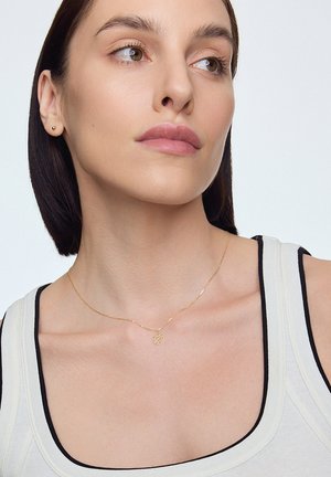 Złoty naszyjnik z delikatnym łańcuszkiem, z okrągłym wisiorkiem o wzorze liścia, noszony na jasnym podkoszulku z ciemnymi akcentami.