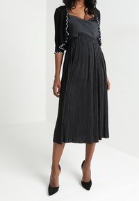 Svart plisserad midiklänning med sidentopp och en kort cardigan med vit volangkant. Slät textur och svävande silhuett.