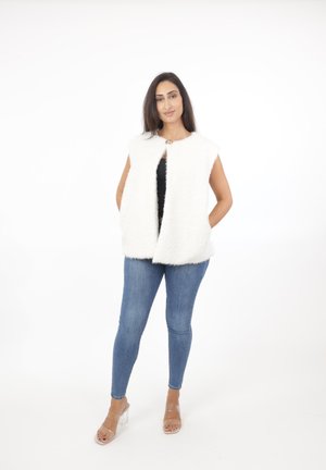 Vrouw staat met een mouwloos wit kunstbontvest, zwarte top, blauwe skinny jeans en doorzichtige sandalen met hoge hakken tegen een witte achtergrond.