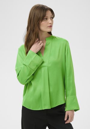 Woman with brown hair wearing a bright green long-sleeve blouse and black pants, looking to the side with hand touching neck.