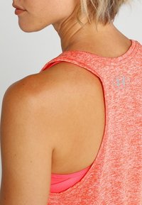 Korall färgad atletisk tanktop med texturerad yta, racerback-utskärning och silverfärgad logodetalj på baksidan. Lagd över en rosa undertröja.