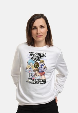Kobieta w białej bluzie z kolorowymi postaciami z Yu-Gi-Oh GX i napisem "Get Your Game On" z przodu, stoi z rękami w kieszeniach.