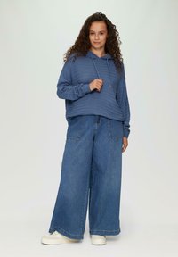 Blauwe hooded sweater met gestructureerde strepen, gecombineerd met wijde denim jeans. Model staat en draagt witte sneakers, tegen een neutrale achtergrond.
