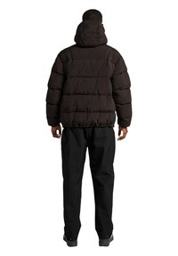 Sort pufferjakke med hætte, der har en quiltet design og elastiske manchetter. Båret med sorte bukser og solide sko, set fra ryggen.