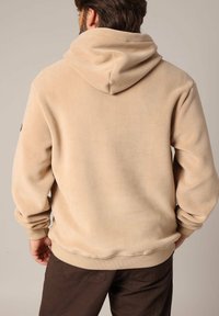 Beige fleece hoodie met een grote capuchon, geribde manchetten en tailleband. Zachte textuur, minimale branding op de mouw, relaxte pasvorm.