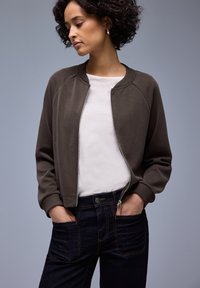 Dunkelgrüne Bomberjacke aus weichem Stoff, mit Reißverschluss vorne, gerippten Bündchen und Kragen, kombiniert mit dunklen Denim-Jeans mit orangefarbenen Nähten.