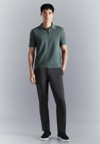 Hombre de pie sobre un fondo gris, vistiendo una camiseta polo verde de manga corta, pantalones negros y zapatillas negras con suelas blancas.