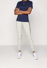 Laivastonsininen lyhythihainen urheilupaita, jossa on Adidas-logo, yhdistetty kerma väriin slim fit -leggingsseihin, joissa on sivuraidat, ja valkoisiin urheilujalkineisiin.
