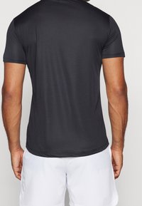 Hombre visto de espaldas, vistiendo una camiseta negra ajustada de manga corta y pantalones cortos blancos holgados, contra un fondo liso.