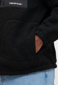 Μαύρο φλις μπουφάν με υφή, διαθέτοντας τσέπη με φερμουάρ στο στήθος και πλαϊνές τσέπες, συμπληρωμένο με τζιν παντελόνι.