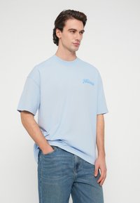 Ανοιχτό μπλε βαμβακερό T-shirt με κοντά μανίκια, στρογγυλή λαιμόκοψη και μικρό κεντημένο λογότυπο στο αριστερό στήθος. Φοριέται με μπλε τζιν τζιν.