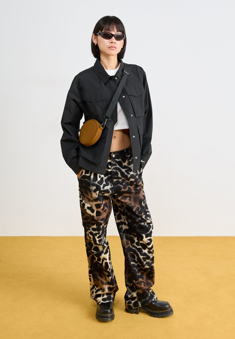 Veste noire à boutons sur un crop top blanc, associé à un pantalon large à imprimé animal, des bottes noires chunky et un sac rond marron.