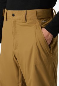 Brune vandtætte bukser lavet af glat stof, med knaplukning, bæltestropper og sidelommer med lynlås. Inkluderer "DRYVENT" branding.