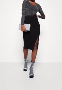 Frau in einem schwarzen gerippten Midirock mit Seitenschlitz, glitzerndem silbernen Oberteil, silberner Clutch und passenden glitzernden silbernen Stiefeletten.