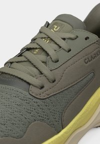 Κοντινή λήψη αθλητικού παπουτσιού σε ελαιοπράσινο χρώμα με υφασμάτινη υφή, κορδόνια στο ίδιο χρώμα, ενισχύσεις από σουέντ και κίτρινη σόλα, με την επωνυμία "Clarks" στη πλευρά.