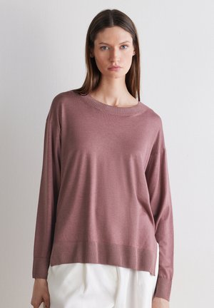 Kobieta o prostych brązowych włosach, ubrana w lawendowy sweter z długim rękawem i białe spodnie, stojąca na tle jasnej, gładkiej ściany.