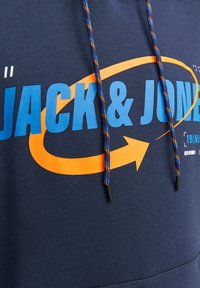 Sudadera azul marino con un gran gráfico en azul y naranja de "JACK & JONES", que presenta un diseño de flecha prominente. Cuerdas para ajustar texturizadas con acentos de color.