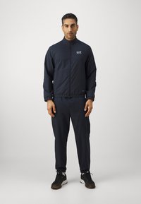 Marine sporttracksuit in textuurstof met een jack met rits en broek met elastische tailleband. Kenmerkt zich door witte branding op het jack. Zwarte sneakers.