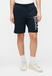 Tmavomodré bavlnené šortky s elastickým pásom a šnúrkou, s bielym nápisom „Emporio Armani“ na boku. Hladká textúra a rovný strih.