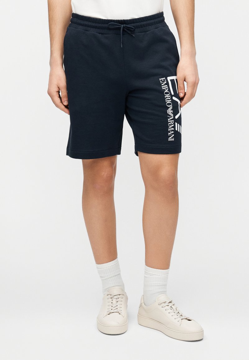 Tmavomodré bavlnené šortky s elastickým pásom a šnúrkou, s bielym nápisom „Emporio Armani“ na boku. Hladká textúra a rovný strih.