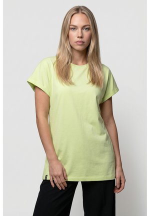 Blonde Frau trägt ein hellgrünes, übergroßes T-Shirt und schwarze Hose, steht vor einem schlichten grauen Hintergrund.