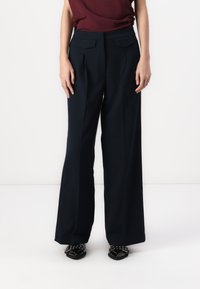 Wide-leg trousers in dark navy fabric with front pleats, a high waist, and side pockets, paired with black patent shoes.
