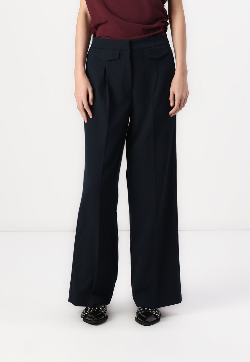 Wide-leg trousers in dark navy fabric with front pleats, a high waist, and side pockets, paired with black patent shoes.
