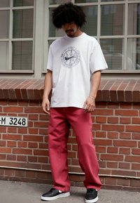 Witte katoenen t-shirt met een grafische print, gecombineerd met rode broeken. Zwarte sneakers maken de outfit compleet. Achtergrond van een stedelijke bakstenen muur.