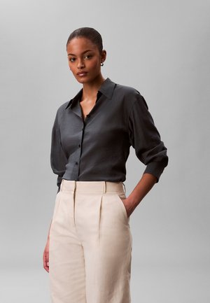 Młoda kobieta z włosami związanymi w kok, ubrana w ciemnoszarą koszulę zapinaną na guziki i beżowe spodnie z wysokim stanem, stojąca z jedną ręką w kieszeni.